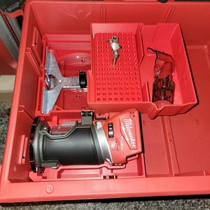 May include: A red Milwaukee FUEL 18V 1/4-inch trim router with a black handle and a clear plastic dust collection shroud is in a red plastic tool box. The router is in a compartment with a grey plastic base plate and a clear plastic dust collection shroud. The tool box has a red plastic tray with a grid of holes for storing bits.
