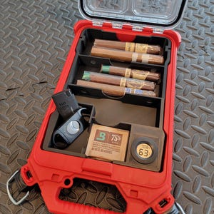May include: A red, open cigar humidor with a clear lid, containing several cigars, a cigar cutter, and a humidity control system. The humidor has a handle and metal loops. The humidity control system reads "75%" and "63".