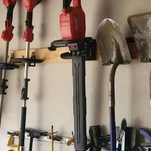 May include: A wall-mounted storage rack with a red and black electric hedge trimmer, two red and black electric weed trimmers, a shovel, and a pair of pruning shears.