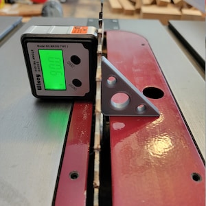 May include: A silver digital angle gauge with a green display reading 0.000. The gauge is labeled "Wiley Digital Angle Gauge" and "Model No. WR300 Type 2". The gauge is sitting on a red surface with a gray plastic triangle.