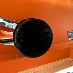 May include: Close-up of a black plastic dust collection port on an orange table saw. The port is round and has a smooth surface.