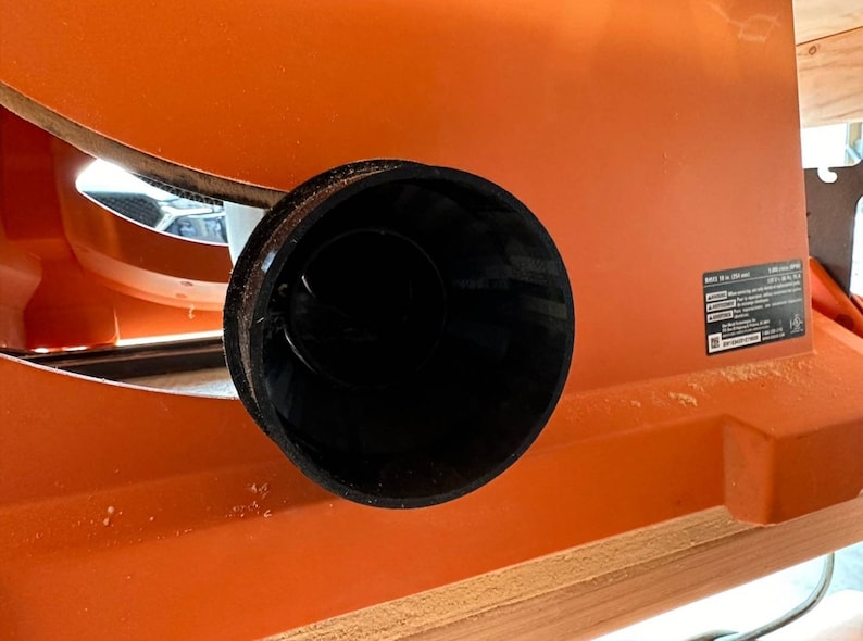 May include: Close-up of a black plastic dust collection port on an orange table saw. The port is round and has a smooth surface.