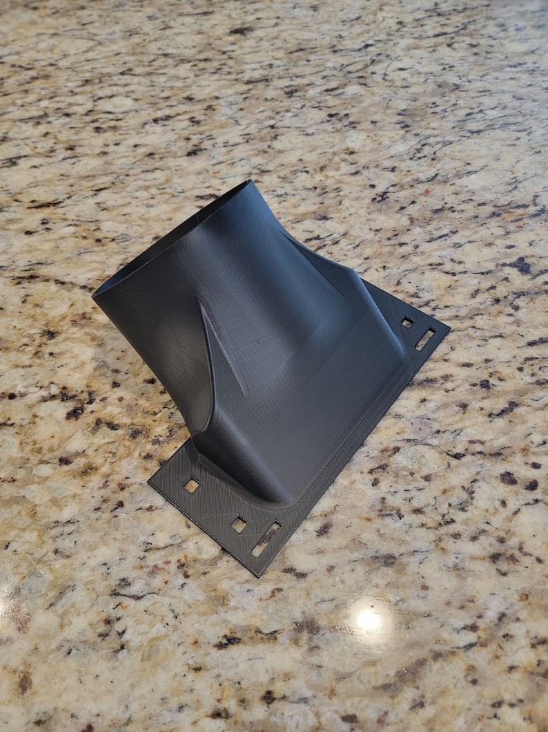 Pode incluir: Um adaptador de duto de ar de pl&aacute;stico preto com uma base retangular e uma abertura redonda. O adaptador tem v&aacute;rios orif&iacute;cios na base.