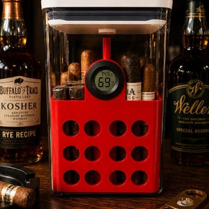 May include: A clear rectangular humidor container with a white lid and a red interior insert featuring twelve circular cutouts. A digital hygrometer is mounted on the front, displaying 74.5 degrees Fahrenheit and 69 percent humidity, with cigars stored inside.
