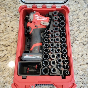 May include: A red Milwaukee M12 FUEL impact driver kit with a clear lid. The kit includes the impact driver, a variety of black sockets, and a battery. The case is open, revealing the tools neatly organized inside.