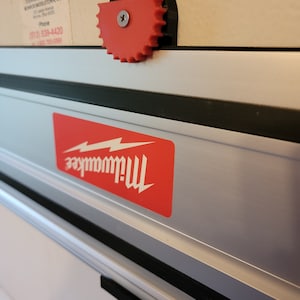 Może przedstawiać: Czerwono-czarne narzędzie elektryczne Milwaukee z czerwoną gałką zębatą. Narzędzie ma czarną i srebrną metalową obudowę oraz czarny plastikowy uchwyt.