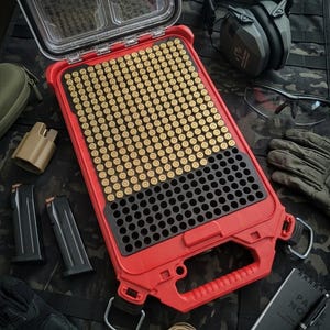 May include: A red and black ammunition storage case with a clear lid, filled with brass-colored cartridges. Additional items include black magazines, ear protection, gloves, and a notepad, all arranged on a camouflage surface.