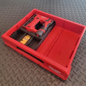 May include: A red Milwaukee Packout tool box containing a red and black nail gun and a yellow and black box of Dewalt nails. The tool box has several compartments. It is on a grey surface.