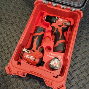 Puede incluir: Un kit de herramientas Milwaukee rojo con tapa transparente. El kit contiene un taladro, un atornillador de impacto y accesorios. Las herramientas son rojas y negras. El texto "Dirty Dog Woodworking" es visible.