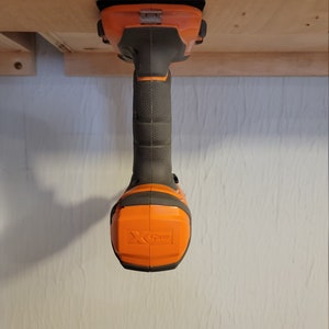 May include: An orange and black cordless drill with a black handle and a black and orange battery pack. The drill is mounted upside down on a wooden surface.