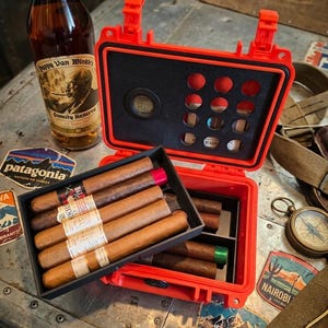 May include: A bright orange hard-shell travel humidor with a digital hygrometer and foam inserts, containing several brown cigars with paper bands. Next to it stands a bottle of Pappy Van Winkle's Family Reserve 15 Years Old bourbon whiskey.