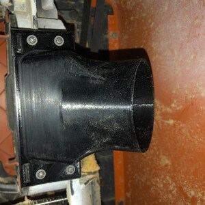 Pode incluir: Porta de coleta de p&oacute; de pl&aacute;stico preto com uma abertura redonda e uma base retangular. A porta &eacute; fixada a uma pe&ccedil;a de maquinaria com parafusos.