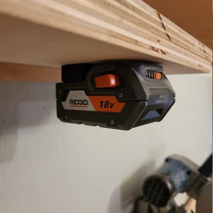May include: A black and orange Ridgid 18V Lithium Ion battery is mounted to a wooden shelf. The battery has a black power button and a black and orange power indicator light.