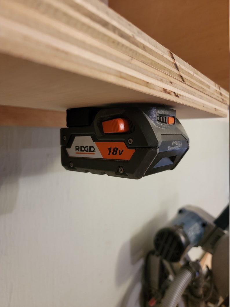 May include: A black and orange Ridgid 18V Lithium Ion battery is mounted to a wooden shelf. The battery has a black power button and a black and orange power indicator light.
