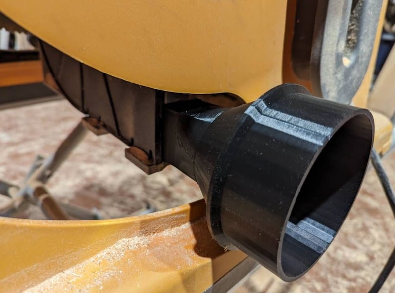 May include: Close-up of a black plastic dust collection port attached to a yellow woodworking machine.