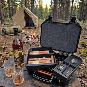 May include: A black hard-shell travel case with foam inserts containing several cigars, a cigar cutter, a lighter, and a round hygrometer. Next to the case are two crystal glasses filled with amber liquid and a bottle of Pappy Van Winkle's Family Reserve.