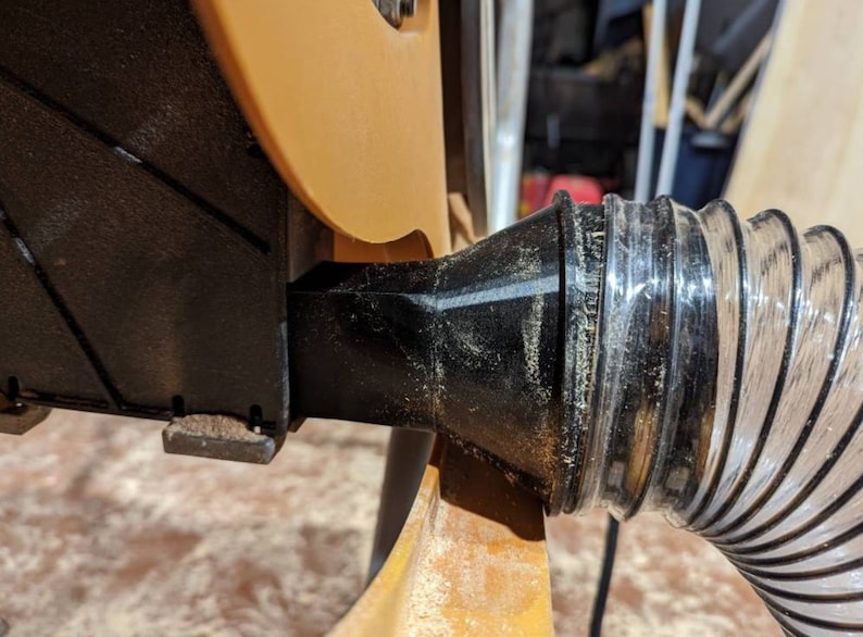May include: Close-up of a black dust collection port on a power tool, connected to a clear plastic hose.