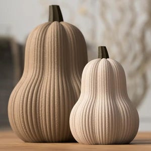 Puede incluir: Dos objetos decorativos con forma de calabaza, uno de color beige claro y otro blanquecino. Ambos tienen detalles de estrías verticales y tallos de color marrón oscuro. Probablemente están hechos de cerámica. El más grande mide unos 25 cm de alto.