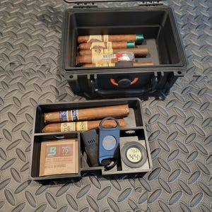 May include: Black, rectangular cigar humidor with cigars, a cutter, and a digital hygrometer. The humidor is open, revealing several cigars with green and gold bands. A small packet with the text "75%" is also visible.