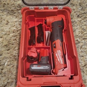 Puede incluir: Un kit de multiherramienta Milwaukee FUEL M12 rojo con accesorios, incluyendo cuchillas y una batería, dentro de una caja roja. La caja tiene una tapa transparente y está abierta, revelando las herramientas. La batería dice "M12 REDLITHIUM HIGH OUTPUT XC5.0."