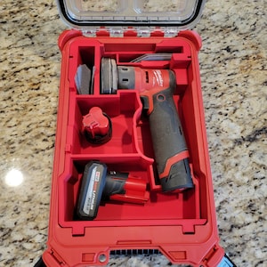 Può includere: Scatola portautensili rossa con una levigatrice del marchio Milwaukee, una batteria e dischi abrasivi al suo interno. La scatola ha un coperchio trasparente e una chiusura su ogni lato.