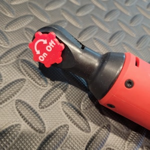 Op de afbeelding: Een rode en zwarte elektrische ratelsleutel met een rode aan/uit-schakelaar. De schakelaar is een rode, stervormige draaiknop met witte tekst en pijlen. De tool is ontworpen voor het aandraaien of losdraaien van bouten en moeren.