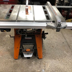 Pode incluir: Uma serra de mesa Ridgid com uma mesa de metal e uma base laranja. A serra tem uma cerca e uma r&eacute;gua de esquadria.
