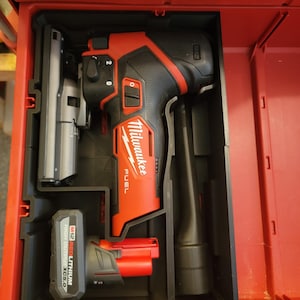 Puede incluir: Una sierra de calar Milwaukee FUEL roja con una hoja negra y gris, un soplador de polvo negro y una batería roja M12 RED LITHIUM XC 5.0 Ah en una caja de herramientas roja.