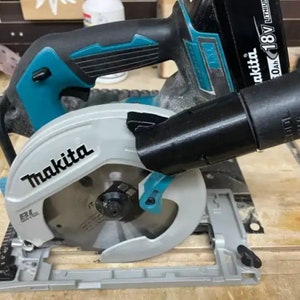 Könnte beinhalten: Eine Makita 18V Akku-Kreissäge mit einem schwarzen und grauen Sägeblattschutz und einem schwarzen Staubabsaugschlauch. Die Säge steht auf einem grauen Sockel mit einer schwarzen und grauen Führungsschiene.