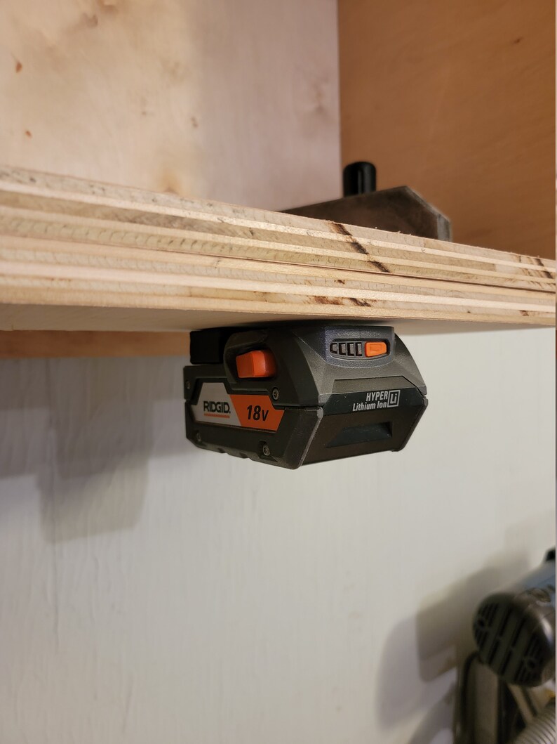 May include: A black and orange 18V lithium-ion battery for a Ridgid power tool. The battery is mounted on a wooden shelf.