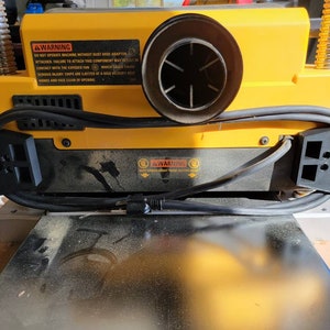 May include: A yellow and black electric planer with a dust collection hose attached. The planer has a warning label that reads "WARNING DO NOT OPERATE MACHINE WITHOUT DUST HOSE ADAPTOR ATTACHED. FAILURE TO ATTACH THIS COMPONENT MAY RESULT IN CONTACT WITH THE EXPOSED FAN. SERIOUS INJURY. CHIPS ARE EJECTED AT A HIGH VELOCITY. KEEP HANDS AND FACE CLEAR OF OPENING."