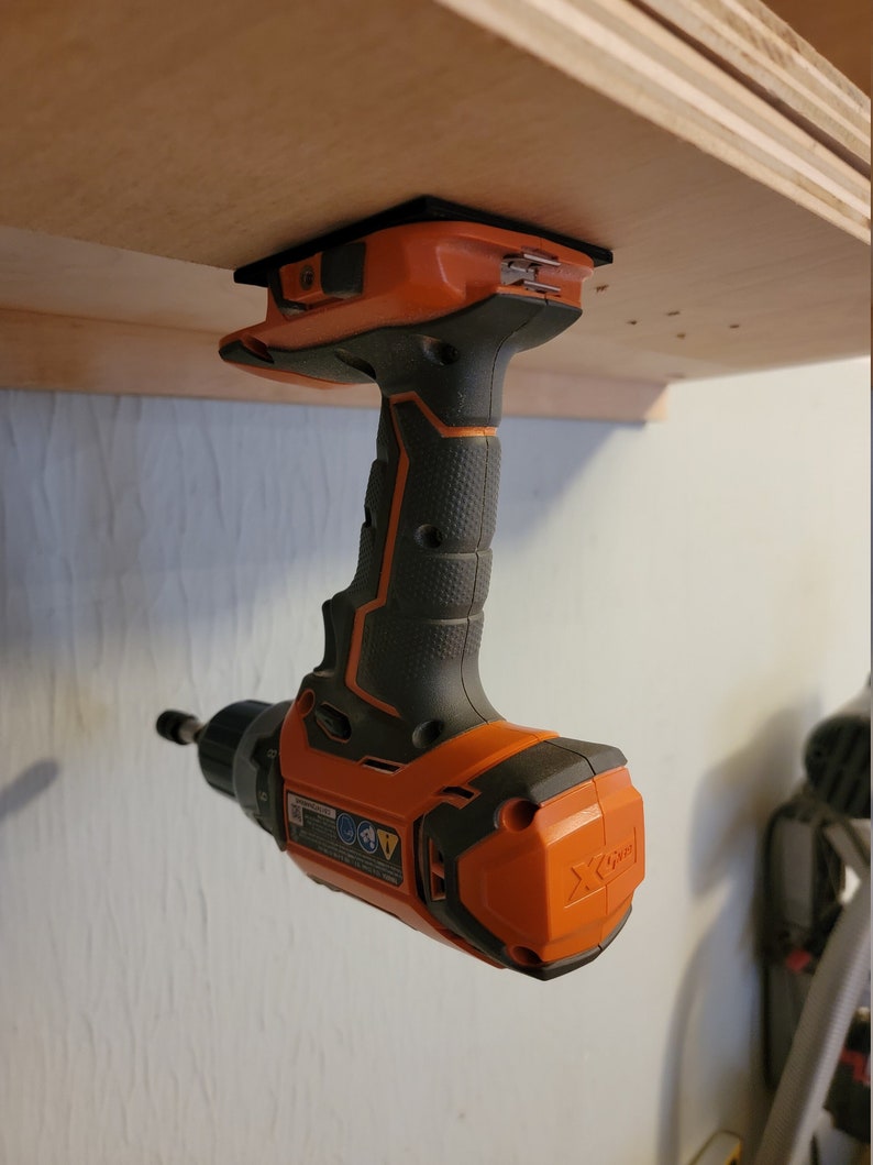 May include: An orange and black cordless drill is mounted upside down on a wooden shelf. The drill has a black handle and a black and orange body. The drill is labelled "X-Drive".