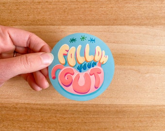 May include: A blue and pink round sticker with the text "FOLLOW YOUR GUT" in a colorful, cartoon font. The gut is depicted as a pink cartoon intestine.