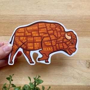May include: A brown and white bison sticker with the outline of the United States, each state labeled with its abbreviation. The bison is standing on all four legs with its head turned to the right.