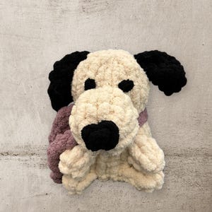 May include: A handmade plush dog toy with a cream-colored body, black ears, and a black nose. The dog has a purple scarf around its neck and is made of a soft, textured material. The toy is designed to look like a friendly dog.