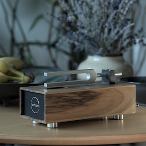 Puede incluir: Un dispositivo de terapia de sonido de madera con un diapasón plateado. El dispositivo tiene un panel negro con la palabra "harmaton". La base de madera se apoya en cuatro patas plateadas. El fondo incluye un jarrón de flores secas y un cuenco de plátanos.