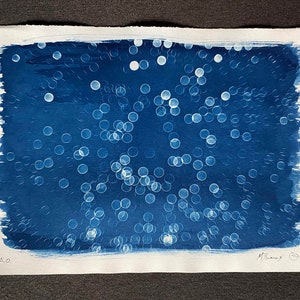 May include: An abstract artwork in blue and white on textured paper. The piece shows a dark blue background with many blurred white circular shapes, like bokeh. The paper edges are rough, and the artwork is signed.