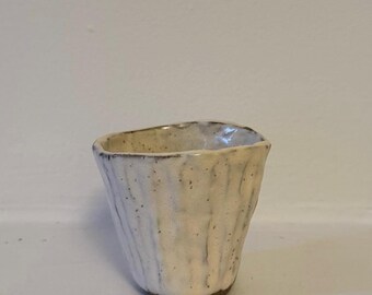 May include: A small, handmade ceramic cup with a textured, off-white exterior. The cup has a slightly irregular shape and a dark brown rim and base. The surface has vertical ridges, giving it a rustic appearance.
