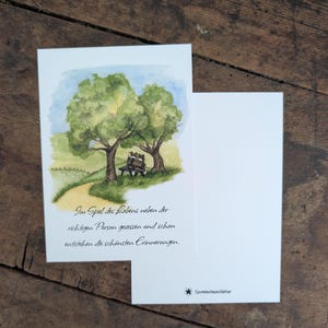 Könnte beinhalten: Eine rechteckige Grußkarte mit einer Aquarellillustration von zwei Hunden, die auf einer Bank unter zwei großen grünen Bäumen auf einer Wiese sitzen. Die Karte enthält den Text: Im Spiel des Lebens neben der richtigen Person gesessen und schon entstehen die schönsten Erinnerungen. Ein kleines Logo mit dem Text Spommelmanufaktur ist unten rechts aufgedruckt.