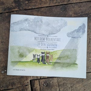 Könnte beinhalten: Ein Kinderbuch mit dem Titel "MIT DEM WOLKENTAXI ZU DEN STERNEN" und dem Text "eine kleine Trauer Fibel". Das Cover zeigt eine Aquarellillustration von Hunden auf einer grünen Wiese unter einem bewölkten Himmel. Der Autor ist "von Nadja & Sylvia".