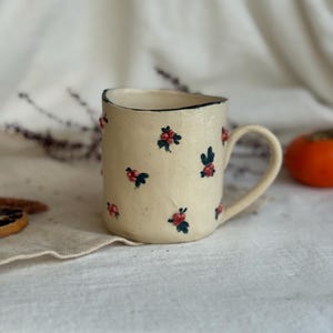 Può includere: Tazza in ceramica color crema con manico, decorata con piccoli motivi floreali rossi e verdi. Il bordo è delineato da una linea blu scuro. La tazza è appoggiata su un tessuto bianco.