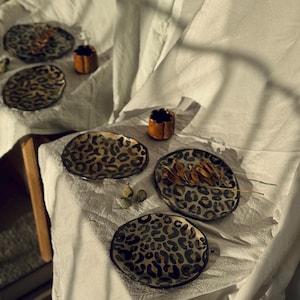 May include: A collection of ceramic plates and small cups with a leopard print design. The plates are round and feature a black and tan animal print. The cups are small, brown, and have a unique shape. The items are arranged on a white cloth.