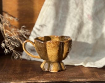 Könnte beinhalten: Eine handgefertigte Keramik-Teetasse mit einem einzigartigen, geriffelten Design. Die Tasse hat eine warme, erdige Farbpalette aus Braun und Gelb mit einem kleinen Henkel. Die Tasse steht auf einem Sockel, was ihren künstlerischen Reiz verstärkt.