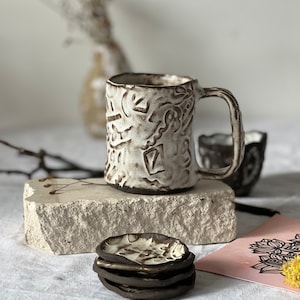 Könnte beinhalten: Eine weiße Keramiktasse mit braunem, strukturiertem Design. Die Tasse steht auf einem Steinuntersetzer und daneben liegen vier kleinere Keramikuntersetzer übereinander.