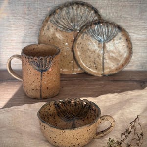 May include: A ceramic tableware set including a mug, a small handled bowl, and two plates. The pieces are speckled with a tan and brown colour scheme, and each has a dark blue floral design. The mug and bowl have a scalloped rim.