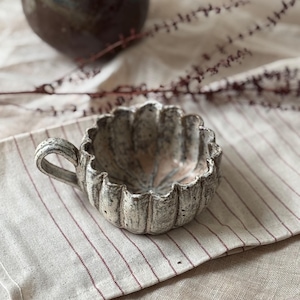 May include: A handmade ceramic teacup with a fluted rim and a small handle. The cup is a neutral colour with a textured, aged appearance. It sits on a striped linen cloth. A dark brown vase is in the background.