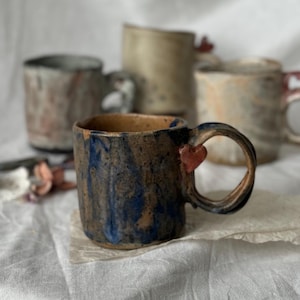 Puede incluir: Colección de tazas de cerámica hechas a mano con diseños únicos. La taza delantera presenta un patrón veteado azul oscuro y marrón con un adorno en forma de corazón en el asa. Otras tazas en el fondo tienen colores y texturas variadas, incluyendo rosa, beige y gris.