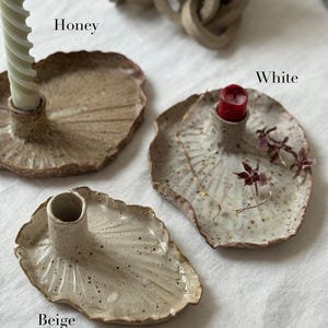 Pode incluir: Uma coleção de castiçais de cerâmica feitos à mão em bege, mel e branco. Cada suporte tem uma forma única de folha com uma taça central para a vela. A imagem inclui uma vela verde e vermelha.
