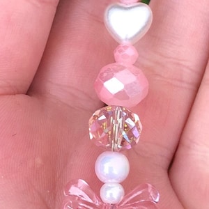 May include: A pink and white beaded charm with a silver clip. The charm features a pink bow, heart-shaped beads, and iridescent beads.