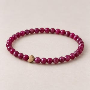 Könnte beinhalten: Ein Perlenarmband mit kleinen, runden, facettierten magentafarbenen Perlen, die in einem Kreis angeordnet sind. Ein einzelner, glatter, metallisch goldfarbener herzförmiger Anhänger ist in die Perlenkette integriert und leicht außermittig platziert.
